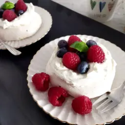 Dwa torty bezowe pavlova z bitą śmietaną, malinami i borówkami na talerzykach.