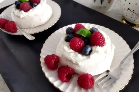 Dwa torty bezowe pavlova z bitą śmietaną, malinami i borówkami na talerzykach.