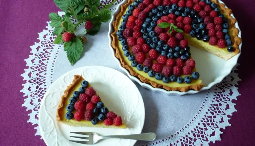 Letnia tarta z kremem budyniowym, ozdobiona malinami i borówkami, kusi apetycznym wyglądem.
