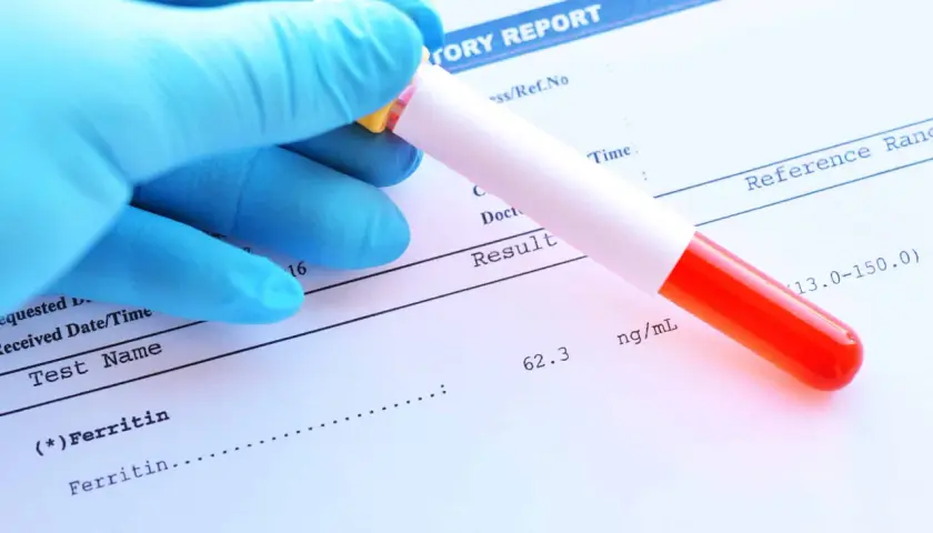 Ręka w niebieskiej rękawiczce trzyma probówkę z krwią nad raportem laboratoryjnym. Wynik ferrytyny to 62.3 ng/ml, co mieści się w normie.