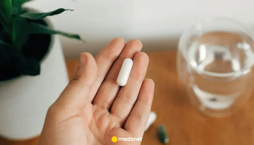 Dłoń trzyma białą kapsułkę. Obok szklanka wody i roślina. Uważaj na nadmiar magnezu objawy.
