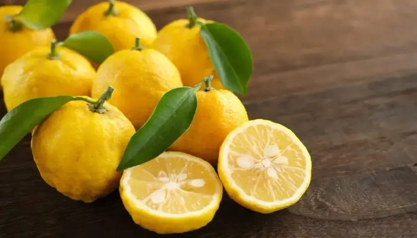 Yuzu co to? Grupa żółtych, pomarszczonych owoców cytrusowych z zielonymi listkami, jeden przekrojony na pół, leżących na drewnianym blacie.